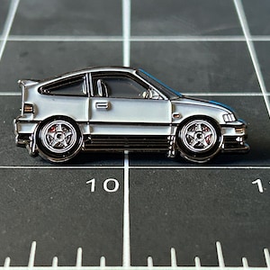 May include: A silver enamel pin of a white Honda Civic del Sol, a two-door sports car with black accents and red brake calipers.