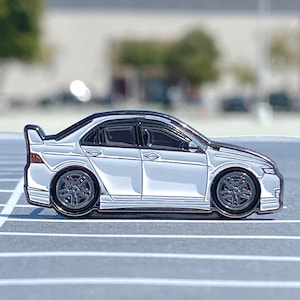 Puede incluir: Un pin de esmalte blanco que representa un coche blanco con detalles negros. El coche es un perfil lateral de una berlina con un alerón en el maletero.