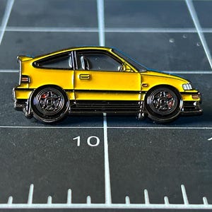 Puede incluir: Un pin de esmalte amarillo de un Honda Civic Del Sol, un modelo de automóvil deportivo popular. El pin está detallado con detalles negros y acentos rojos en las ruedas.
