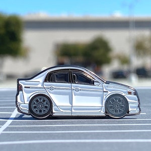 May include: A white enamel pin of a silver sedan with black accents. The car is in profile view and has black wheels.