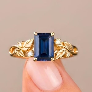 May include: A gold ring featuring a central emerald-cut dark blue sapphire. The band is designed with sculpted gold leaves and small, clear round-cut accent stones embedded within the foliage on both sides of the primary blue gemstone.