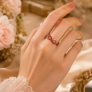 May include: A rose gold ring featuring an infinity symbol design adorned with small, round, vibrant red gemstones set along the top curve of the band. The metal has a polished finish, and the ring is worn on a finger, showcasing the detailed craftsmanship.