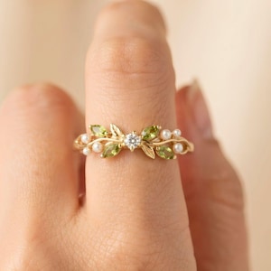 May include: A gold ring featuring a central round clear gemstone flanked by marquise-cut green peridot stones and small white pearls arranged in a leaf-like pattern along the band. The metal band has a delicate, vine-inspired design.