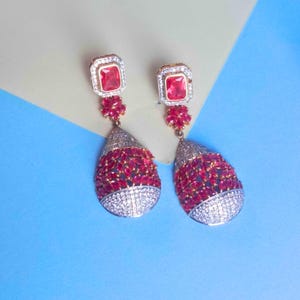 May include: A pair of elegant drop earrings. Each earring features a square, red gemstone at the top, a small floral accent, and a teardrop-shaped pendant encrusted with red and white gemstones. The earrings have a gold-colored setting.
