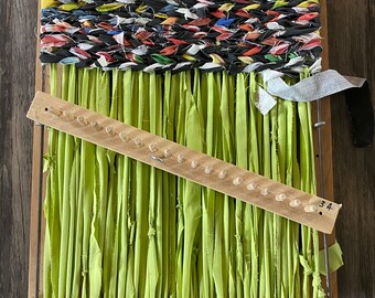 May include: A wooden loom with green fabric strips woven through the warp threads. The loom has a wooden shuttle and a metal hook. The fabric strips are tied to the bottom of the loom. The loom is on a wooden floor.