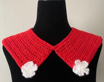 May include: A handmade, red crochet collar with two white flower accents. The collar is displayed on a black mannequin. The crochet work is visible, showcasing the texture and craftsmanship of the piece. A unique accessory.