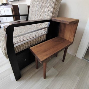 May include: A small wooden side table with a natural wood finish. The table has a rectangular top and two legs. The table is next to a brown and white striped armchair.
