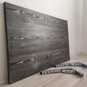 K&ouml;nnte beinhalten: Eine gro&szlig;e, rechteckige Holz-Tischplatte mit dunkelgrauer Beize, die markante Maserungsmuster aufweist. Zwei Metall-Klapphalterungen sind darunter positioniert und bereit zur Installation. Die Tischplatte lehnt an einer wei&szlig;en Wand.