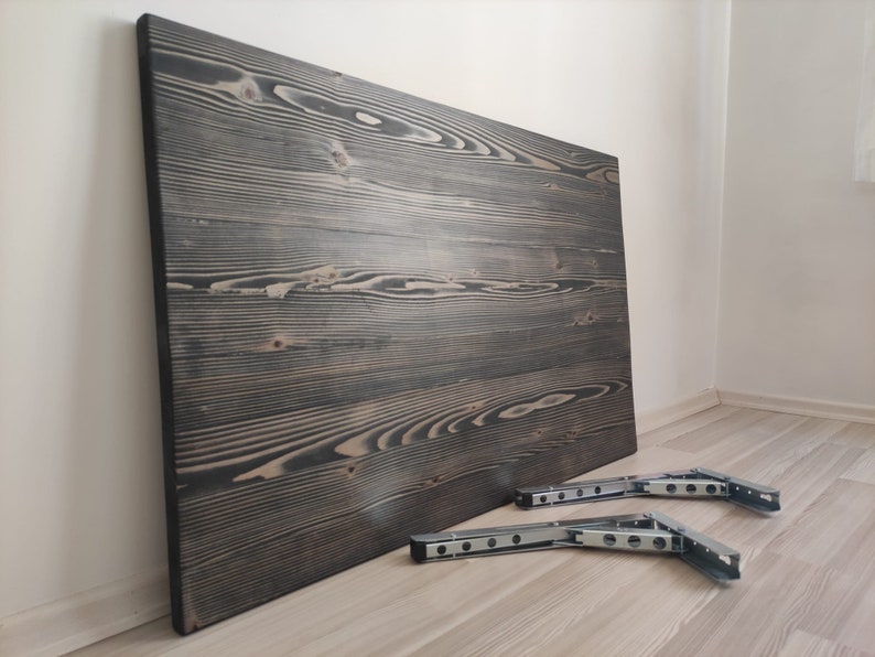 K&ouml;nnte beinhalten: Eine gro&szlig;e, rechteckige Holz-Tischplatte mit dunkelgrauer Beize, die markante Maserungsmuster aufweist. Zwei Metall-Klapphalterungen sind darunter positioniert und bereit zur Installation. Die Tischplatte lehnt an einer wei&szlig;en Wand.