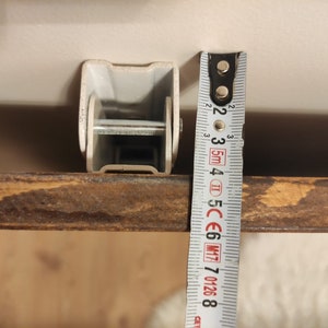K&ouml;nnte beinhalten: Nahaufnahme einer Metallhalterung mit einem Ma&szlig;band daneben. Die Halterung ist hellgrau und rechteckig. Das Ma&szlig;band zeigt ein Ma&szlig; von ca. 5 cm an.