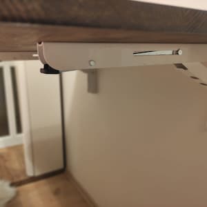 K&ouml;nnte beinhalten: Eine wei&szlig;e Klapptischhalterung ist an einer beigefarbenen Wand montiert und tr&auml;gt eine Holzplatte. Die Halterung hat eine Reihe von L&ouml;chern und einen Verriegelungsmechanismus. Der Tisch ist so konzipiert, dass er bei Nichtgebrauch zusammengeklappt werden kann.