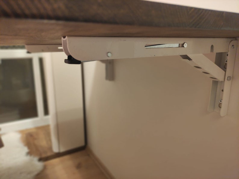 K&ouml;nnte beinhalten: Eine wei&szlig;e Klapptischhalterung ist an einer beigefarbenen Wand montiert und tr&auml;gt eine Holzplatte. Die Halterung hat eine Reihe von L&ouml;chern und einen Verriegelungsmechanismus. Der Tisch ist so konzipiert, dass er bei Nichtgebrauch zusammengeklappt werden kann.
