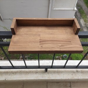 May include: A wooden balcony shelf with a raised back, stained in a dark brown color. The shelf is mounted on a black metal railing.