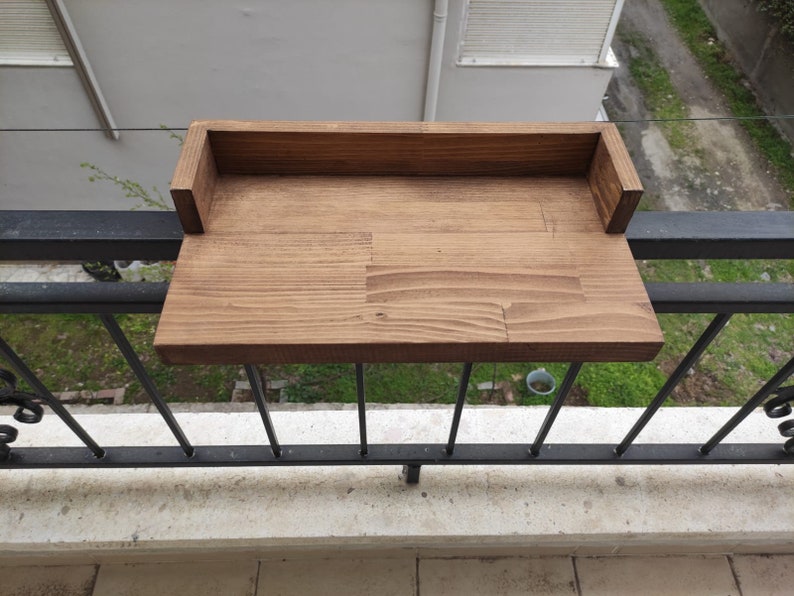 May include: A wooden balcony shelf with a raised back, stained in a dark brown color. The shelf is mounted on a black metal railing.