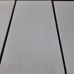 K&ouml;nnte beinhalten: Drei wei&szlig;e Holzpaneele mit einer hellen Holzmaserung. Jedes Paneel hat ein kleines, dunkles Loch in der N&auml;he der Mitte. Die Paneele sind nebeneinander angeordnet, durch eine dunkle Linie getrennt. Oberhalb der Paneele ist eine dunkelbraune Holzoberfl&auml;che zu sehen.