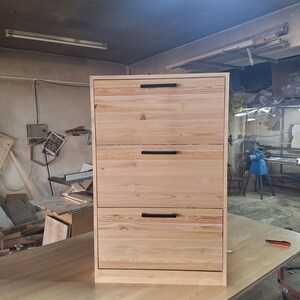 May include: A three-drawer dresser made of light wood with black metal handles. The dresser is standing on a light wood surface.
