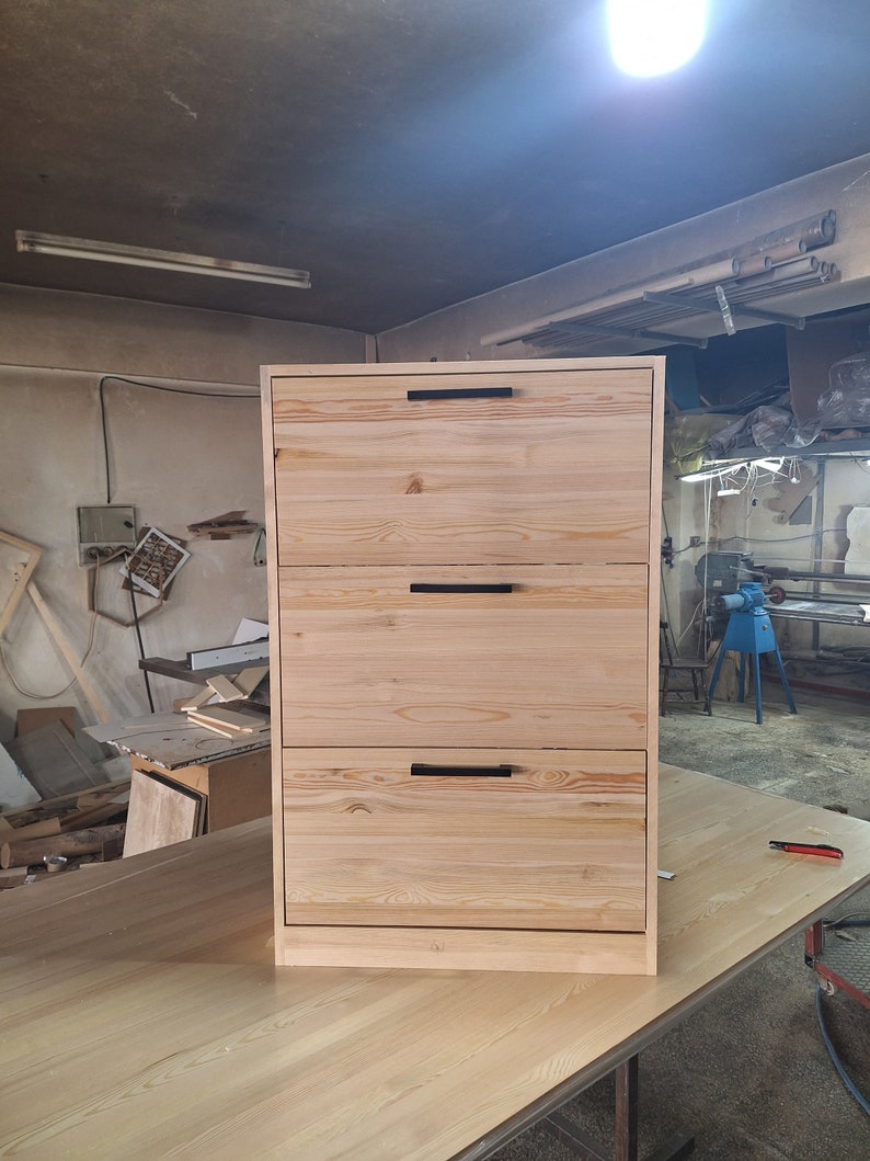 May include: A three-drawer dresser made of light wood with black metal handles. The dresser is standing on a light wood surface.
