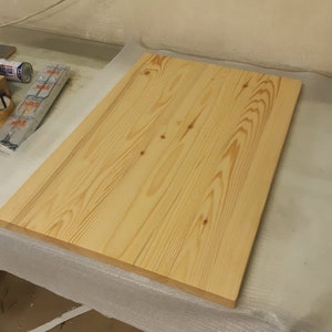K&ouml;nnte beinhalten: Eine rechteckige, unfertige Holzplatte mit einer hellen, nat&uuml;rlichen Holzmaserung. Das Holz hat sichtbare Knoten und eine glatte Oberfl&auml;che. Die Tischplatte ist wahrscheinlich f&uuml;r ein Schreibtisch- oder Tischprojekt.
