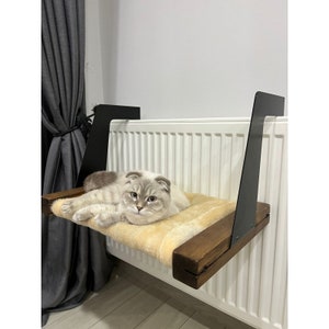 Könnte beinhalten: Ein Katzenbett aus Holz und Stoff, das mit schwarzen Metallhalterungen an einem weißen Heizkörper befestigt ist. Das Bett ist beige und ein Kätzchen liegt darauf.