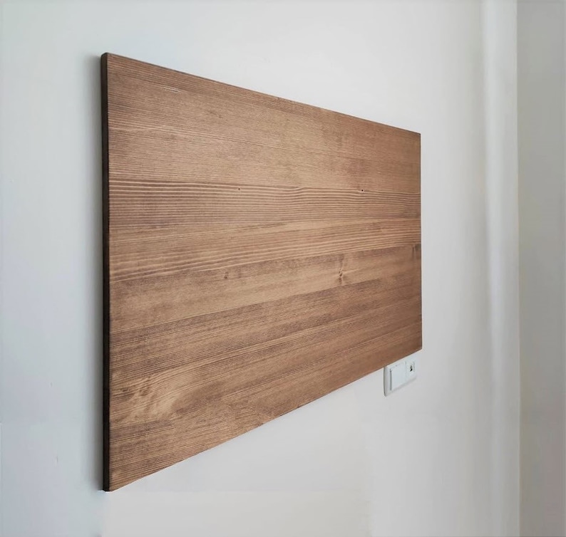 K&ouml;nnte beinhalten: Eine rechteckige Holztafel mit nat&uuml;rlicher brauner Oberfl&auml;che, an einer wei&szlig;en Wand befestigt. Die Holzmaserung ist sichtbar, und die Tafel ist schr&auml;g positioniert, wodurch ihre Textur und Farbe zur Geltung kommen. Die Tafel ist wahrscheinlich ein dekoratives Element oder ein M&ouml;belst&uuml;ck.