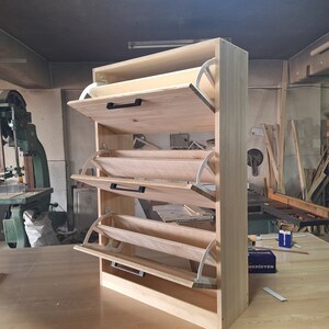 May include: A wooden shoe rack with four shelves and a hinged door on each shelf. The shelves are made of light wood and the doors are white. The shoe rack is standing on a wooden floor.
