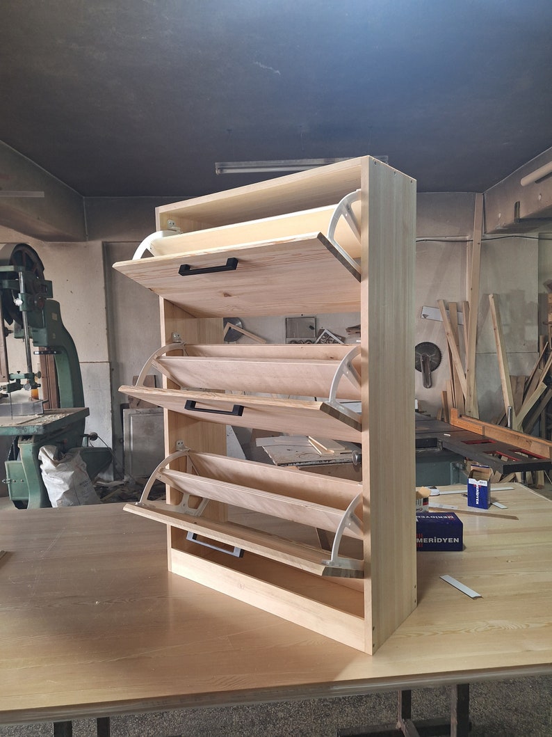 May include: A wooden shoe rack with four shelves and a hinged door on each shelf. The shelves are made of light wood and the doors are white. The shoe rack is standing on a wooden floor.