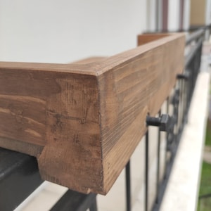 May include: Close-up of a wooden balcony railing with a dark brown stain. The railing is attached to a black metal railing with screws.