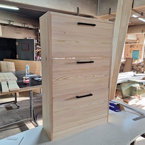 May include: A three-drawer wooden cabinet with black handles. The cabinet is made of light wood and has a simple design.
