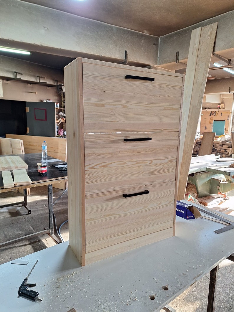 May include: A three-drawer wooden cabinet with black handles. The cabinet is made of light wood and has a simple design.