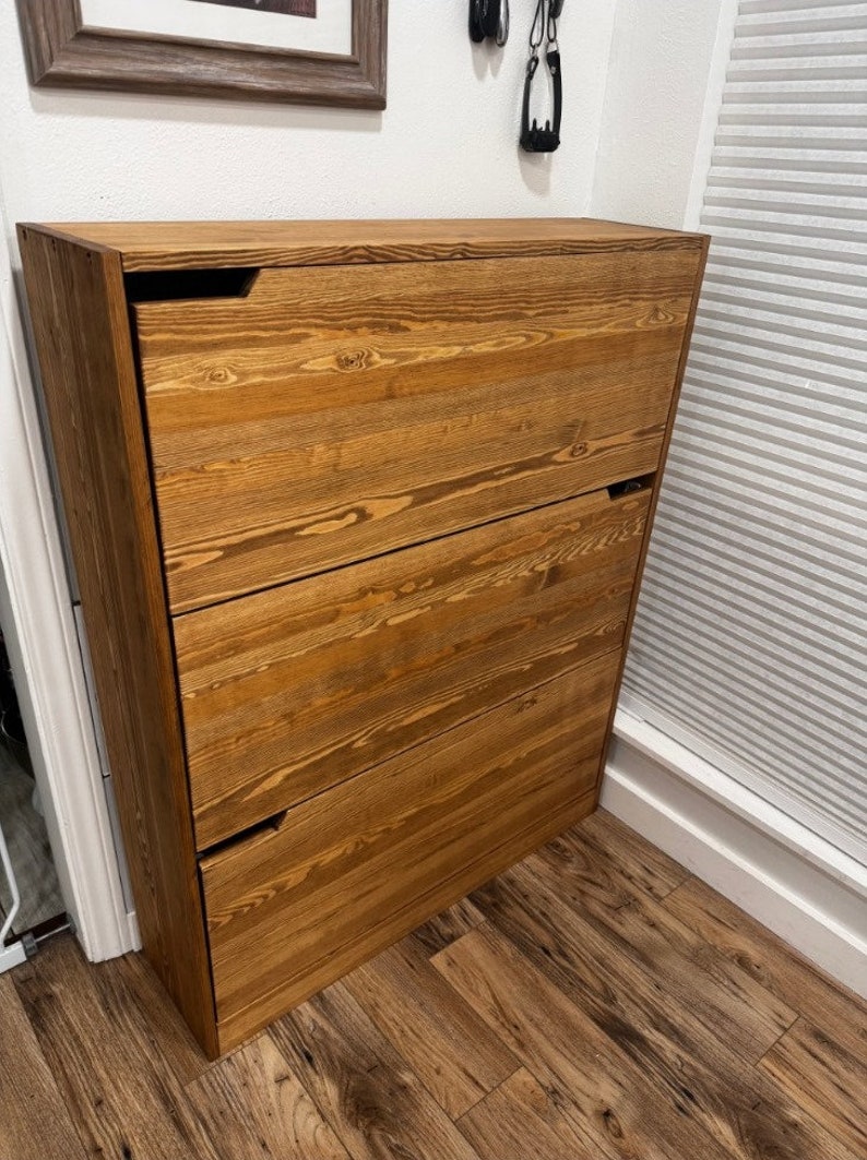 May include: A wooden shoe storage cabinet with three shelves and a natural wood finish. The cabinet has a simple design with no visible hardware.