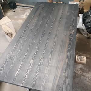 K&ouml;nnte beinhalten: Ein gro&szlig;er, rechteckiger Holztisch mit dunkelgrauer Beize. Die Holzmaserung ist sichtbar, mit helleren Streifen, die &uuml;ber die Oberfl&auml;che verlaufen. Der Tisch wird von Metallbeinen getragen. Die Abmessungen des Tisches sind nicht sichtbar.