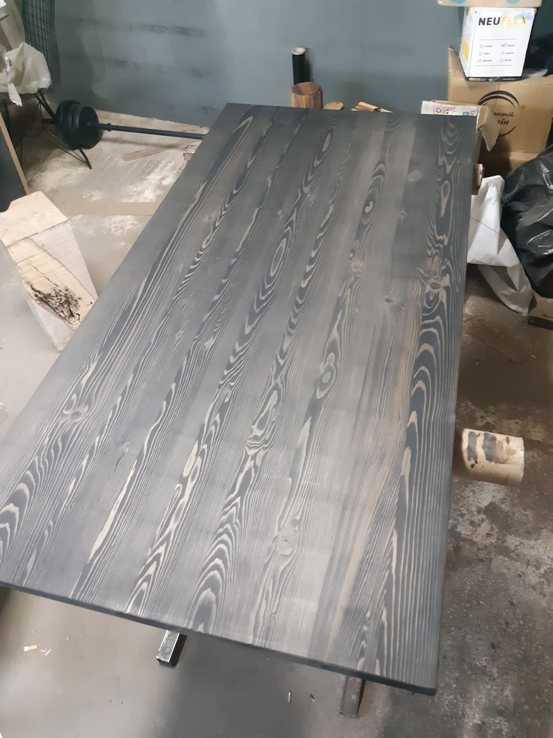 K&ouml;nnte beinhalten: Ein gro&szlig;er, rechteckiger Holztisch mit dunkelgrauer Beize. Die Holzmaserung ist sichtbar, mit helleren Streifen, die &uuml;ber die Oberfl&auml;che verlaufen. Der Tisch wird von Metallbeinen getragen. Die Abmessungen des Tisches sind nicht sichtbar.