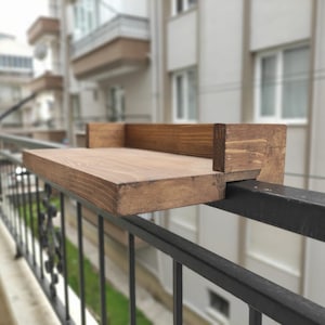 May include: A wooden shelf attached to a black metal railing. The shelf is made of unfinished wood and has a natural brown color.