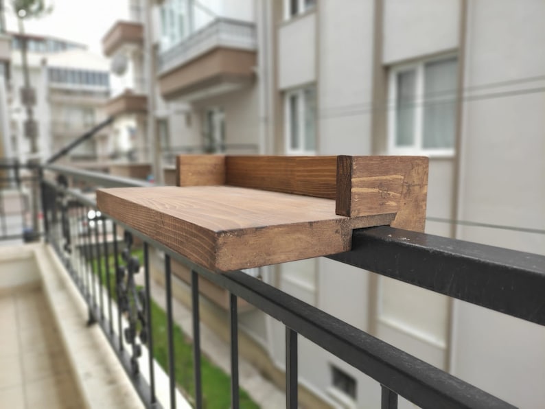 May include: A wooden shelf attached to a black metal railing. The shelf is made of unfinished wood and has a natural brown color.
