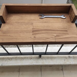 May include: A wooden shelf attached to a black metal railing. The shelf is stained a dark brown color and has a natural wood grain. A silver wrench is lying on the shelf.