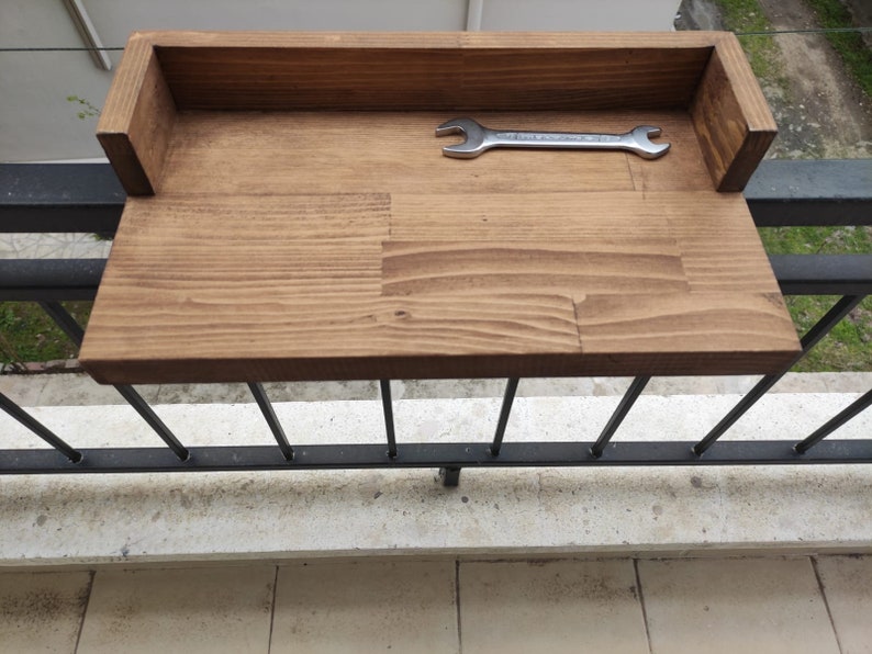 May include: A wooden shelf attached to a black metal railing. The shelf is stained a dark brown color and has a natural wood grain. A silver wrench is lying on the shelf.