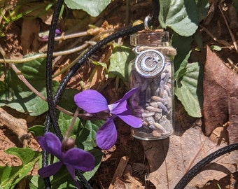 May include: A glass bottle necklace with a crescent moon charm and lavender inside. The necklace is laying on a bed of green leaves and brown dirt.