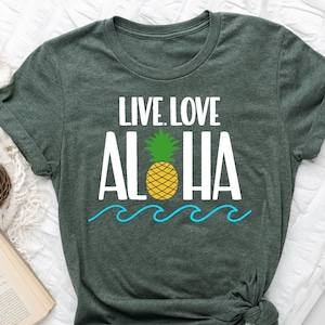May include: Heather forest green t-shirt with the words "LIVE LOVE ALOHA" in white, a yellow pineapple, and blue waves. The shirt is displayed on a white surface with a coffee cup, a book, and a hat.