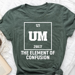 May include: A dark green t-shirt with the text "UM The Element of Confusion" in a white box. The shirt has a crew neck and short sleeves. The shirt is displayed on a white surface with a book, a clock, and a coffee cup.