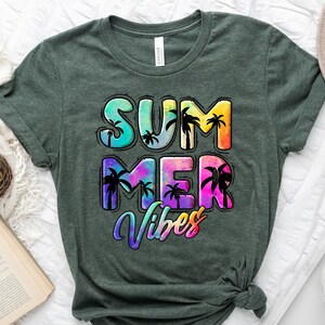 May include: Heather forest green t-shirt with the words "SUMMER VIBES" in a colorful, tropical design. The text features palm tree silhouettes and a tie-dye effect. A coffee cup, a book, and a hat are in the background.