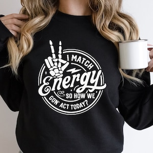Op de afbeelding: Zwart sweatshirt met een wit grafisch ontwerp met een skelethand die een peace-teken maakt en de tekst "I Match Energy So How We Gon' Act Today?"