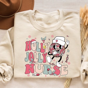 Op de afbeelding: Een cr&egrave;mekleurige sweatshirt met de woorden "HOLLY JOLLY NURSE" in een retro lettertype. Het ontwerp toont een discobal-personage met een verpleegstersmuts. Hulst- en sneeuwvlokaccenten maken het feestelijke ontwerp compleet.