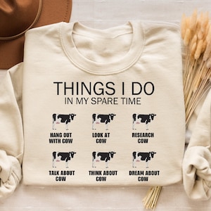 May include: A beige sweatshirt with the text "THINGS I DO IN MY SPARE TIME" and a series of black and white cow illustrations with text below each cow: "HANG OUT WITH COW", "LOOK AT COW", "RESEARCH COW", "TALK ABOUT COW", "THINK ABOUT COW", and "DREAM ABOUT COW".