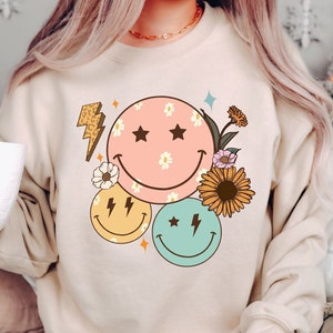 May include: A beige sweatshirt with a graphic design of three smiley faces with stars and lightning bolts. The smiley faces are surrounded by flowers and a lightning bolt.