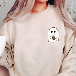 Puede incluir: Sudadera beige con un peque&ntilde;o dise&ntilde;o de fantasma blanco sosteniendo una taza de caf&eacute;. El fantasma tiene ojos negros y est&aacute; delineado en negro. La sudadera tiene mangas largas y cuello redondo. En la imagen se sostiene una taza blanca.
