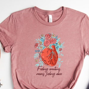 May include: A heather mauve t-shirt featuring a graphic of a heart with flowers and the text "Feeling emotions, means feeling alive".