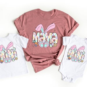 Pu&ograve; includere: Un set di tre magliette con un design di coniglio pasquale. Le magliette dicono "Mama" e "Mini" in un font colorato e a motivi. Le magliette sono bianche con orecchie di coniglio rosa e piedi di coniglio blu.