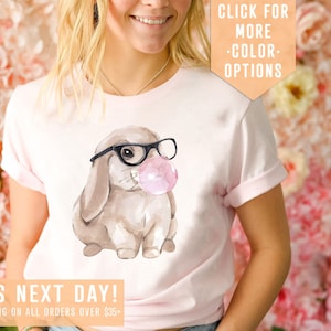 May include: Pink t-shirt with a watercolor illustration of a brown bunny wearing black glasses and blowing a pink bubble. The text "SHIPS NEXT DAY! FREE SHIPPING ON ALL ORDERS OVER $35+" is at the bottom of the shirt. The text "CLICK FOR MORE -COLOR- OPTIONS" is in the top right corner of the image.