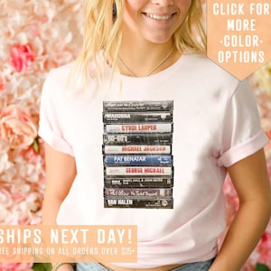 May include: A white t-shirt with a stack of vintage cassette tapes featuring artists like Madonna, Cyndi Lauper, and Michael Jackson. The text "SHIPS NEXT DAY! FREE SHIPPING ON ALL ORDERS OVER $35+" is at the bottom of the image. The text "CLICK FOR MORE -COLOR- OPTIONS" is at the top of the image.