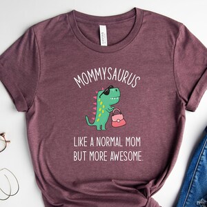 May include: Heather maroon t-shirt with the text "MOMMYSAURUS" and a cartoon dinosaur wearing sunglasses and carrying a purse. The text below reads "LIKE A NORMAL MOM BUT MORE AWESOME."